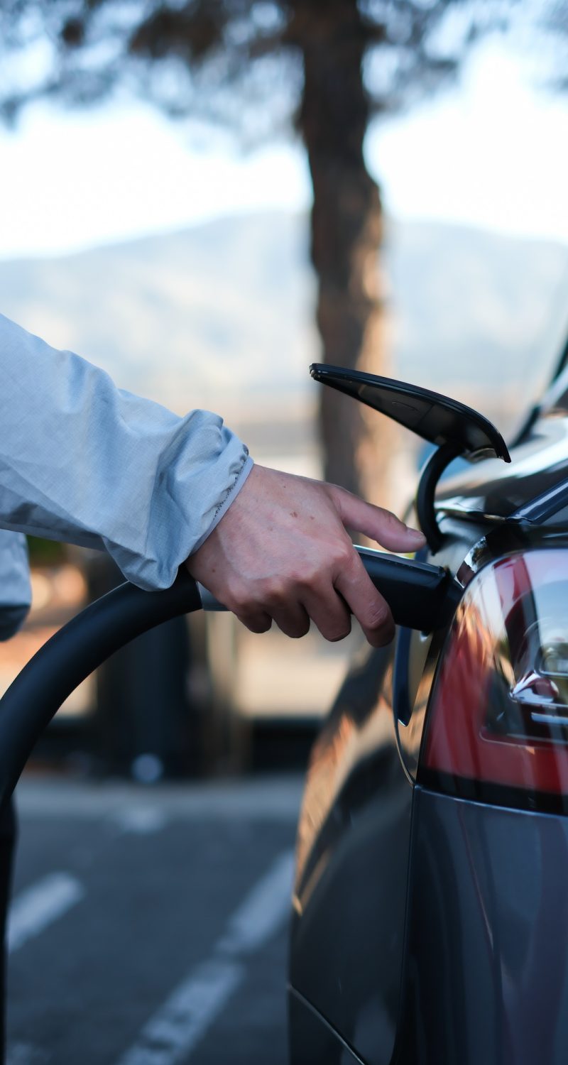 Hand charging EV plug to electric vehicle with natural background for traveling.